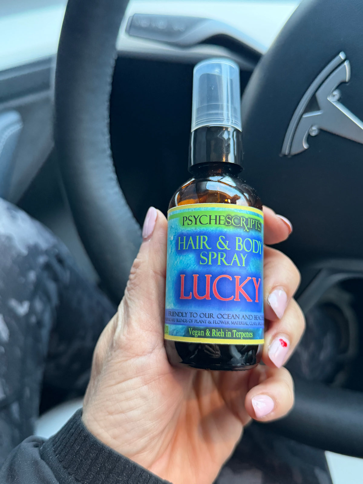 A hand holding a bottle of Psychescrupps 'Lucky' Hair & Body Spray inside a car, with the steering wheel and dashboard visible in the background.