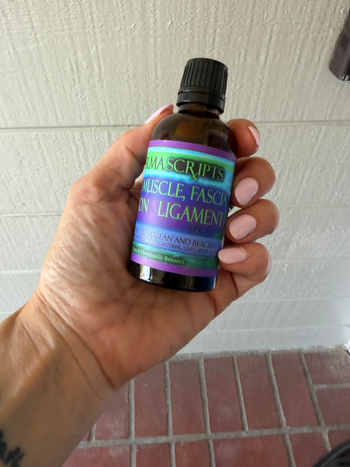 A hand holding a small amber glass bottle with a black cap and a purple, green, and blue label. The label reads 'Elixir Scripts Muscle, Fascia, and Ligament Support'.