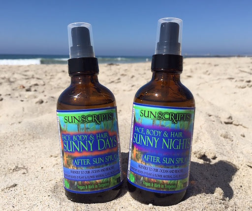 Two amber spray bottles of Sunscriptis Face, Body & Hair 'Sunny Days' After Sun Spray and 'Sunny Nights' After Sun Spray standing on a sandy beach with the ocean and blue sky in the background.